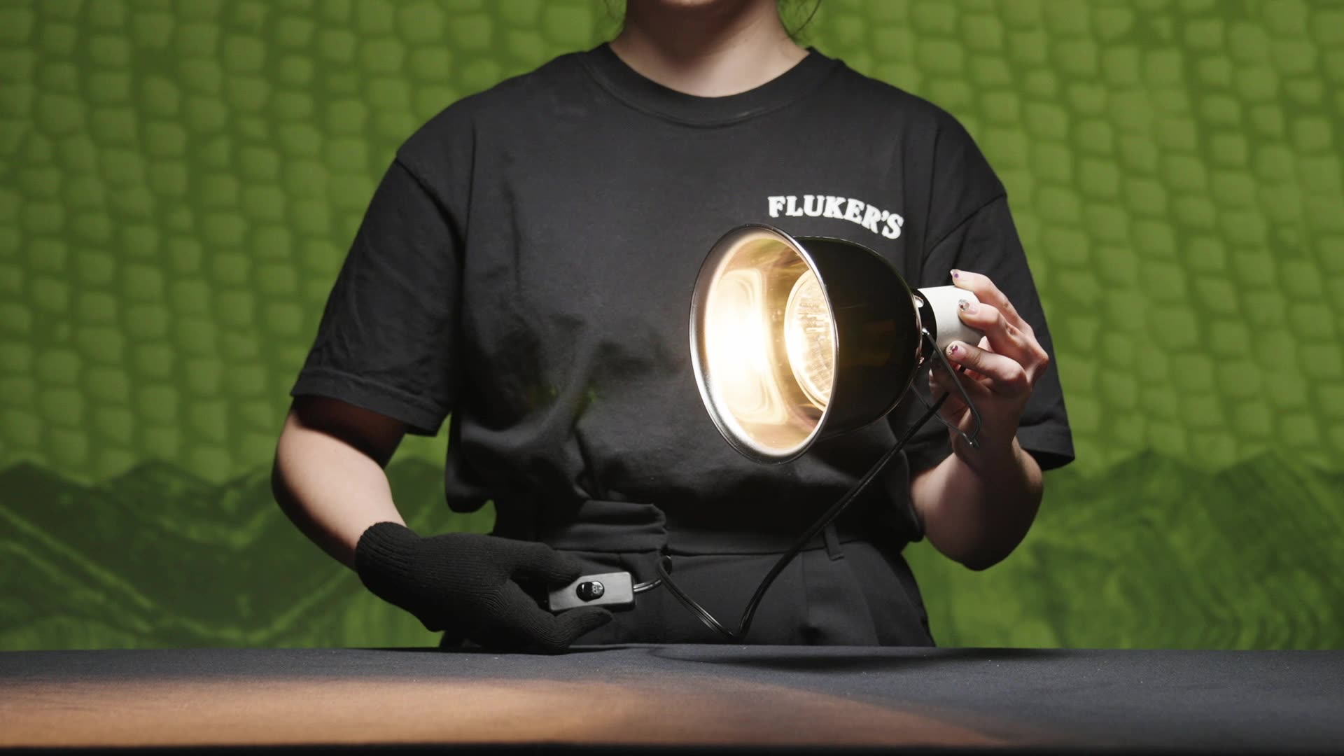 Person in Black Fluker's t-shirt holding Fluker's Deep Doe lamp wearing gloves and showcasing on/off switch with the lamp on